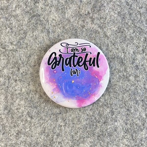 May include: A white button with a purple and pink watercolor background and gold glitter. The button says "I am so grateful for:" in black cursive lettering.