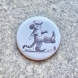 May include: A white button with a black and white illustration of a cat wearing a cowboy hat and boots. The cat is walking with one boot in the air. The background is a light gray with a cactus.