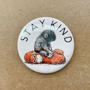 May include: A white button with the words "STAY KIND" in black. The button features an illustration of a gray monkey hugging an orange stuffed animal. The button is round and has a glossy finish.