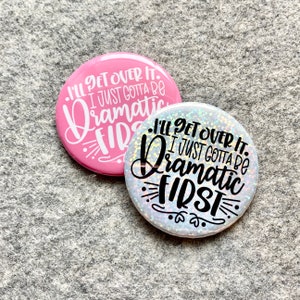 May include: Two round buttons with the text "I'll get over it. I just gotta be dramatic first." The button on the left is pink with white text. The button on the right is silver with black text.