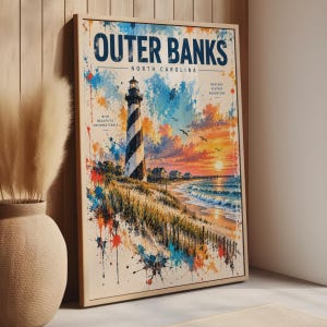 May include: A framed vertical wall art print featuring a watercolour-style illustration of the Outer Banks, North Carolina lighthouse. The scene includes a striped lighthouse, coastal dunes, ocean waves, and a sunset, with text accents throughout.