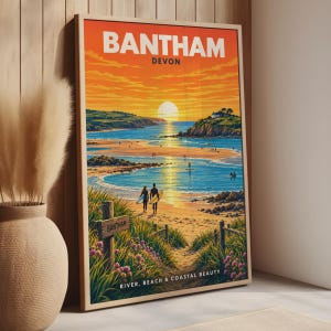 Pode incluir: Um cartaz de viagem emoldurado de Bantham, Devon, com um pôr do sol sobre uma praia de areia e água azul. O texto diz BANTHAM DEVON e RIVER, BEACH & COASTAL BEAUTY. Duas figuras com pranchas de surf estão na costa perto de dunas e flores.