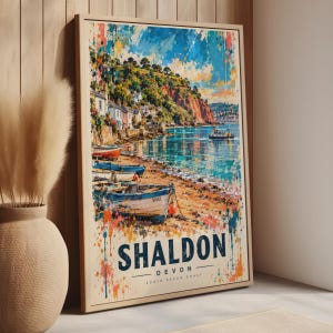 May include: A framed vertical wall art print featuring a watercolour-style coastal scene of Shaldon, Devon. The image depicts colourful boats on a sandy shore, a calm blue sea, and hillside buildings. The text SHALDON DEVON SOUTH DEVON COAST is printed.