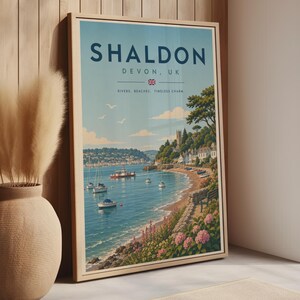 Puede incluir: Un póster de viaje vertical enmarcado que muestra una escena costera de Shaldon, Devon, Reino Unido. La ilustración representa una bahía azul con veleros, un pequeño ferry, una playa y una colina con edificios. El texto dice SHALDON DEVON, UK.