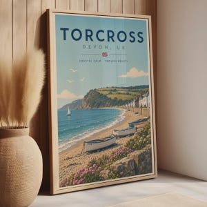 May include: A framed travel poster featuring a coastal scene of Torcross, Devon, UK. The print displays a sandy beach with small boats, a calm blue sea, green hills, and white buildings. The text reads TORCROSS DEVON, UK and COASTAL CALM. TIMELESS BEAUTY.