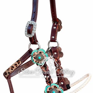 May include: A brown leather horse headcollar featuring leopard print accents, braided leather details, and silver-toned buckles. The headcollar is decorated with flower-shaped ornaments in turquoise and leopard print, and includes a white rope lead.