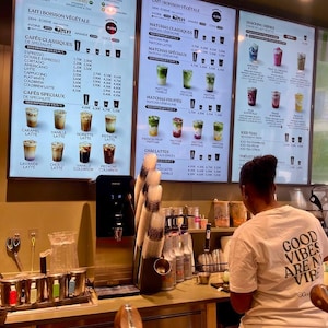以下が含まれることがあります： カフェの内観で、メニューボードには飲み物のオプションが表示されています。メニューにはコーヒー、抹茶、アイスティーが含まれています。 「GOOD VIBES ARE M VIBE」という文字が入った白いTシャツを着た人がカウンターの後ろに立っています。
