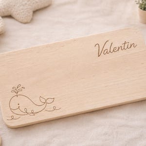 May include: A rectangular light-colored wooden board with rounded corners. The surface features a laser-engraved line drawing of a whale spouting water in the bottom left corner and the name Valentin in cursive script in the top right corner.