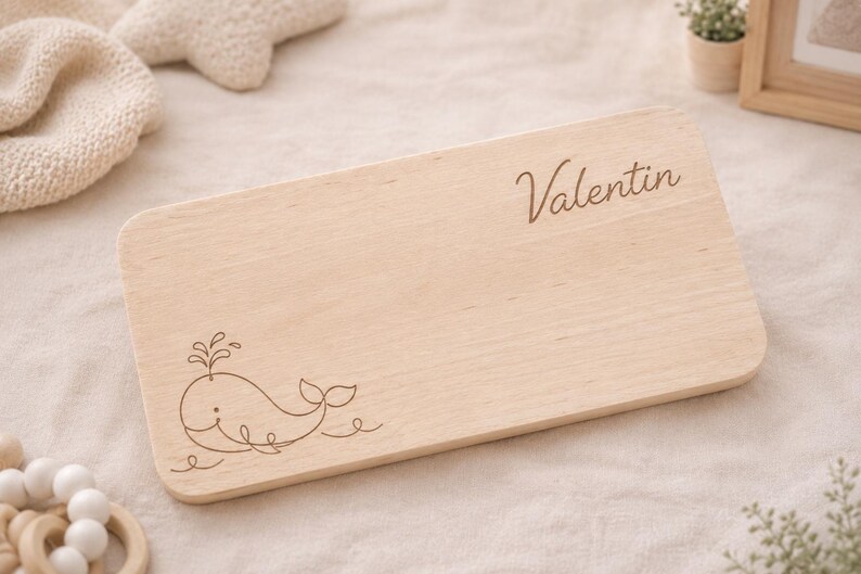 May include: A rectangular light-colored wooden board with rounded corners. The surface features a laser-engraved line drawing of a whale spouting water in the bottom left corner and the name Valentin in cursive script in the top right corner.