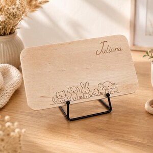 May include: A rectangular light-colored wooden plaque resting on a black metal stand. The plaque features the engraved name Juliana in cursive script and a line art illustration of a cat, dog, rabbit, bird, and elephant along the bottom edge.