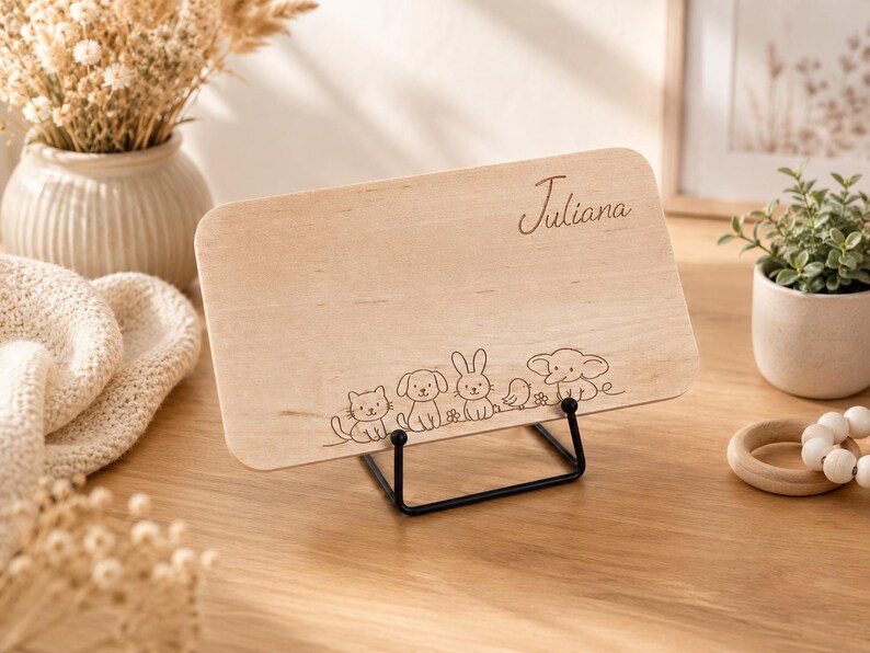 May include: A rectangular light-colored wooden plaque resting on a black metal stand. The plaque features the engraved name Juliana in cursive script and a line art illustration of a cat, dog, rabbit, bird, and elephant along the bottom edge.