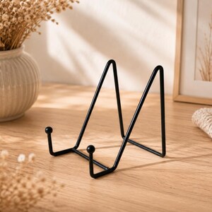 May include: A black metal display stand featuring a triangular frame with two front-facing prongs topped with small spheres. The stand is constructed from thin, smooth wire and is designed to hold items upright in a stable, angled position.