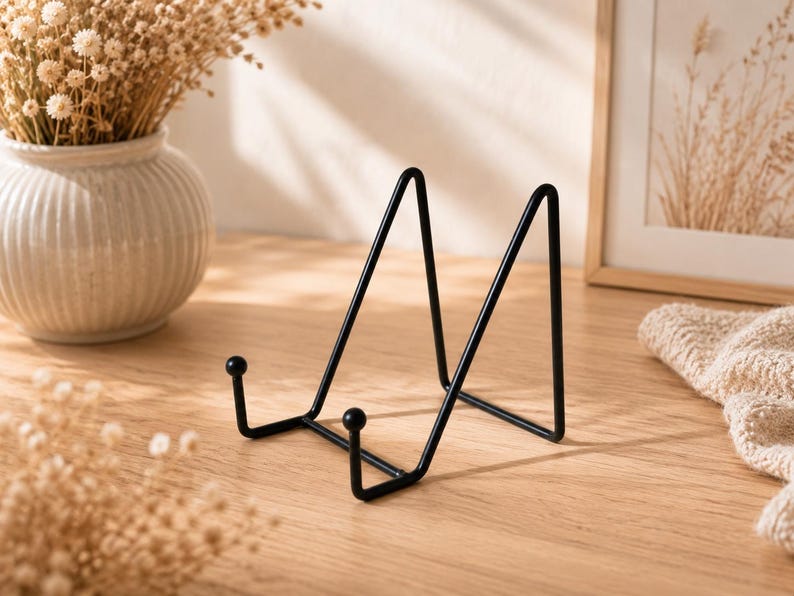 May include: A black metal display stand featuring a triangular frame with two front-facing prongs topped with small spheres. The stand is constructed from thin, smooth wire and is designed to hold items upright in a stable, angled position.