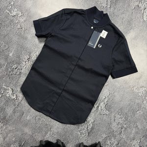 Könnte beinhalten: Schwarzes Polohemd mit kurzen Ärmeln, strukturiertem Muster und kleinem weißem Logo auf der Brust. Das Hemd hat einen Knopfkragen und ein angebrachtes Etikett. Das Hemd wird auf einer grauen Oberfläche präsentiert.