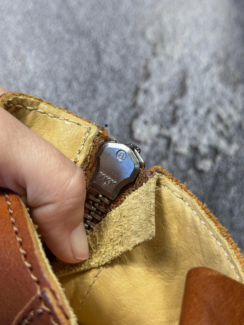 May include: Close-up of a brown leather boot with a silver zipper. The zipper is partially open, revealing the interior lining. The zipper pull is marked with the letters "YKK". The boot has visible stitching and a worn appearance.