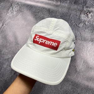 Pode incluir: Boné de beisebol branco com um patch retangular vermelho que diz "Supreme" em letras brancas. O boné tem uma aba curva e uma coroa estruturada. Feito de um material leve.