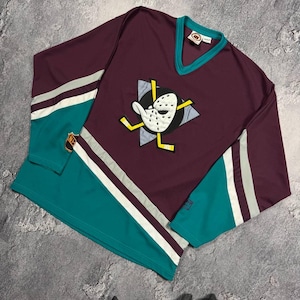 May include: A burgundy, teal and white hockey jersey with a V-neck. The jersey has a duck-masked character logo on the front. The sleeves feature white and grey stripes. A vintage sports top.