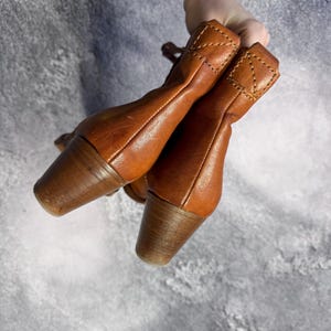 May include: A pair of brown leather ankle boots with wooden heels. The boots have a classic design with a rounded toe and a stitched detail at the top of the boot. The heels are approximately 2 inches (5 cm) high.