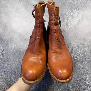 May include: A pair of brown leather ankle boots with a zipper closure and a rounded toe. The boots have a distressed look and are held up by a hand. The boots are approximately 6 inches tall.