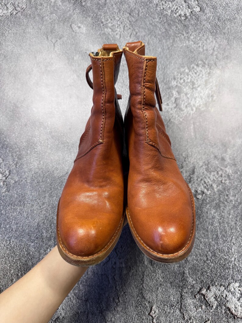 May include: A pair of brown leather ankle boots with a zipper closure and a rounded toe. The boots have a distressed look and are held up by a hand. The boots are approximately 6 inches tall.