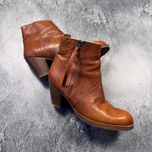 May include: A pair of brown leather ankle boots with a zipper closure and a stacked heel. The boots have a rounded toe and a pull tab. The leather has a slightly distressed appearance.