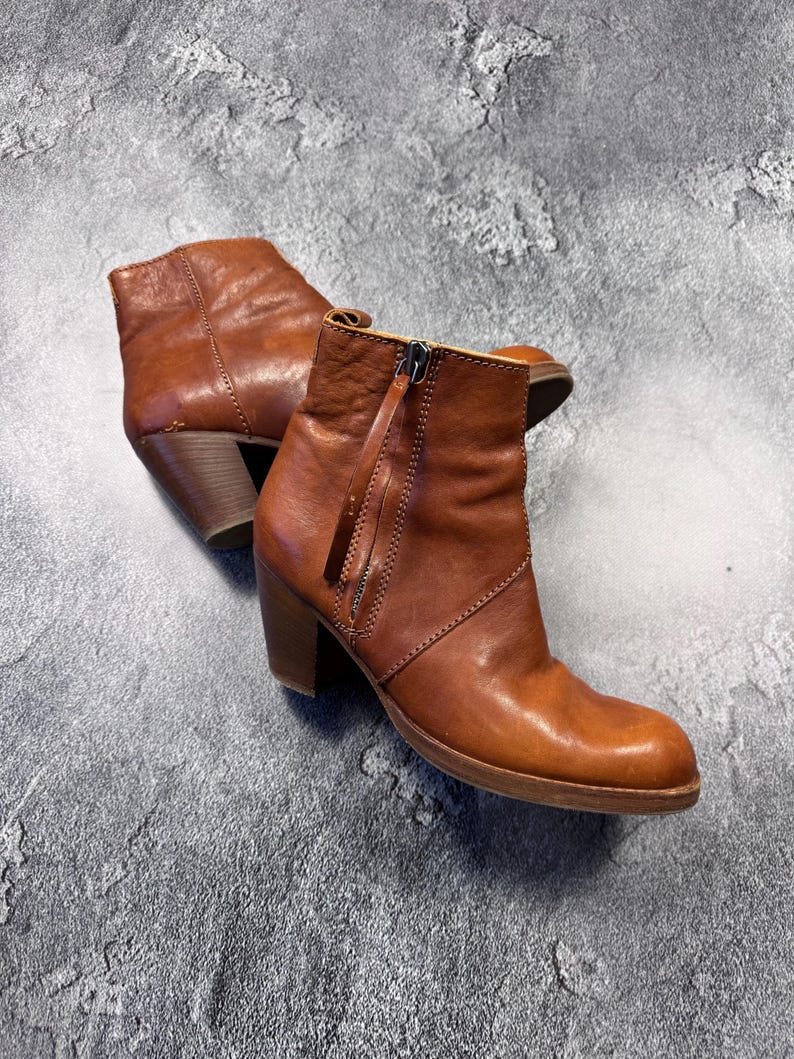 May include: A pair of brown leather ankle boots with a zipper closure and a stacked heel. The boots have a rounded toe and a pull tab. The leather has a slightly distressed appearance.