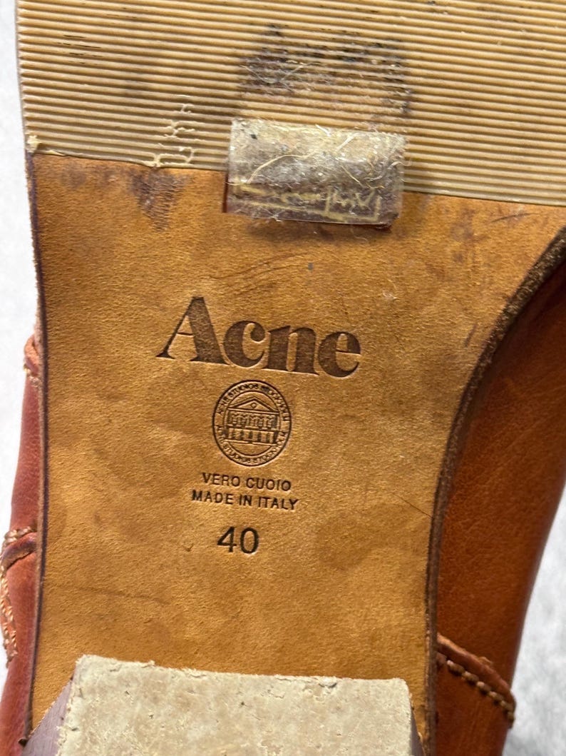 May include: Close-up of a brown leather shoe sole with the brand name "Acne" embossed. The sole also features the text "VERO CUOIO MADE IN ITALY" and the number "40".