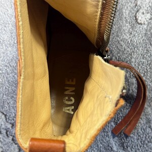 May include: A close-up view inside a brown leather boot. The interior is tan with the word "ACNE" stamped in gold. A zipper runs along the side, and a leather pull tab is visible. The boot has a worn, vintage appearance.