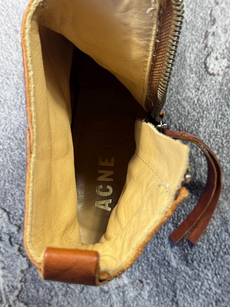 May include: A close-up view inside a brown leather boot. The interior is tan with the word "ACNE" stamped in gold. A zipper runs along the side, and a leather pull tab is visible. The boot has a worn, vintage appearance.