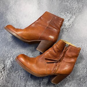 May include: A pair of brown leather ankle boots with a side zipper and a stacked heel. The boots have a rounded toe and a distressed finish, suggesting a vintage or worn look. The boots are set against a gray background.