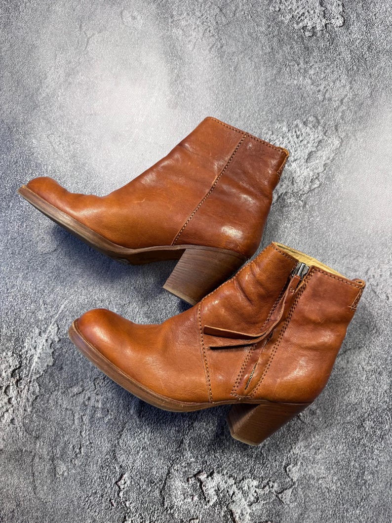 May include: A pair of brown leather ankle boots with a side zipper and a stacked heel. The boots have a rounded toe and a distressed finish, suggesting a vintage or worn look. The boots are set against a gray background.