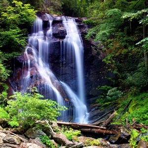 Op de afbeelding: Een schilderachtige waterval stort naar beneden over een donkere rotswand, omgeven door weelderig groen gebladerte. Het water stroomt over de rotsen en creëert een mistig effect. Omgevallen boomstammen en grote rotsen bevinden zich aan de basis, waardoor een natuurlijk landschap ontstaat.