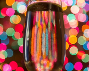 May include: A glass of champagne with colourful lights reflected in the liquid. The lights are out of focus and create a festive atmosphere.