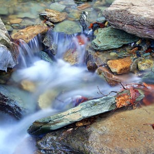 May include: A scenic image of a small waterfall cascading over colorful rocks. The water flows smoothly, creating a blurred effect, with hints of ice and a few fallen leaves. The rocks are various sizes and colors.