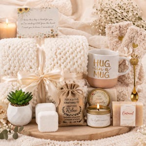 May include: A gift basket with a cream-colored blanket tied with a satin ribbon, a mug with "HUG in a MUG" text, a candle, soap, and a small succulent. Includes a card with text and a small burlap bag.