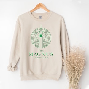 Könnte beinhalten: Beiges Sweatshirt mit einem grünen Grafikdesign, das ein kreisförmiges Design mit einem Auge und architektonischen Elementen zeigt. Darunter steht in Grün "THE MAGNUS ARCHIVES". Das Sweatshirt hängt an einem Holzkleiderbügel.