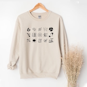 Könnte beinhalten: Beiges Sweatshirt mit einer Anordnung schwarzer grafischer Symbole, darunter eine Flamme, ein Auge und ein Totenkopf. Die Symbole sind in Reihen und Spalten auf der Vorderseite angeordnet. Das Sweatshirt hängt an einem Holzkleiderbügel.
