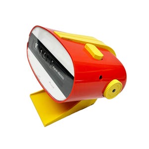 May include: A vintage red and yellow digital-electric flip clock. The clock has a curved red body with yellow accents and a yellow base. The clock face displays the time with black numbers on a white background.