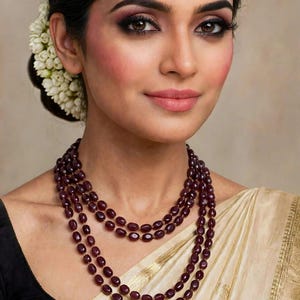 May include: A multi-strand necklace featuring polished, deep burgundy oval beads. The necklace consists of three distinct layers of beads that drape around the neck, creating a layered appearance with a consistent dark red color throughout.
