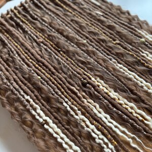 May include: A set of brown and white synthetic dreadlocks with gold and white beads. The dreadlocks are arranged in rows on a wooden surface.