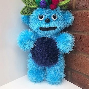 Puede incluir: Una criatura de peluche azul hecha a mano con una panza azul oscuro y una boca sonriente. Tiene ojos blancos y un adorno para la cabeza decorativo con hojas verdes, bayas moradas y bayas rojas. El peluche está sobre una superficie blanca.