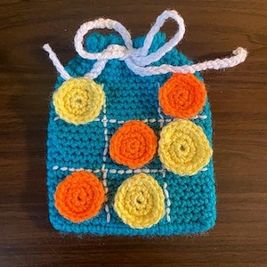 May include: A handmade, turquoise crochet Tic-Tac-Toe game bag with a white string closure. The game board is stitched with a grid and features orange and yellow crochet game pieces. A fun, portable game.