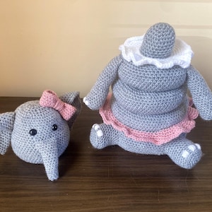 Puede incluir: Dos juguetes de elefante de ganchillo gris. Uno es una cabeza con un lazo rosa. El otro es un elefante apilado con un cuello blanco y una falda rosa. Los juguetes están hechos a mano y están sobre una superficie de madera.
