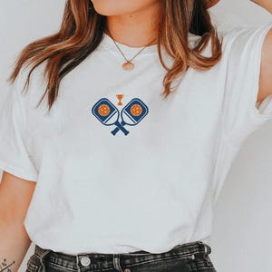 May include: White t-shirt featuring a blue and orange pickleball design. The graphic includes crossed paddles, a pickleball, and a trophy. The shirt is made of a soft material and has a classic crew neck.