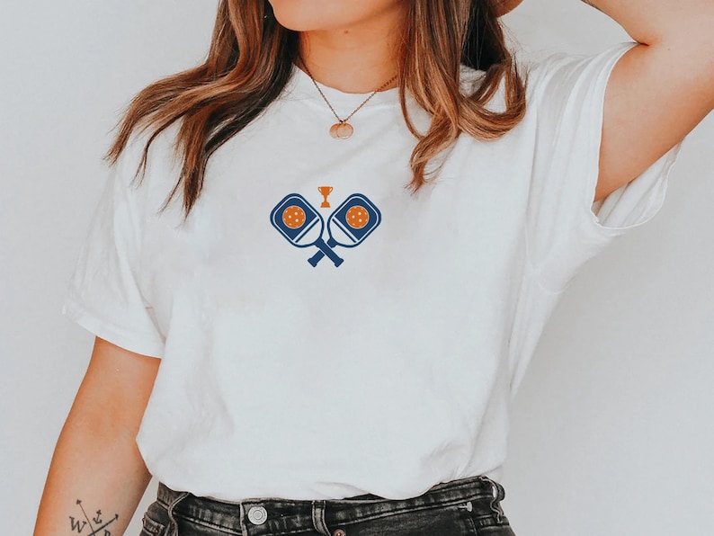 May include: White t-shirt featuring a blue and orange pickleball design. The graphic includes crossed paddles, a pickleball, and a trophy. The shirt is made of a soft material and has a classic crew neck.