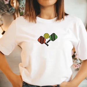 May include: White t-shirt featuring a colorful pickleball design. The graphic includes a yellow pickleball and two paddles, one red and one green. The paddles have black handles and vertical stripes.