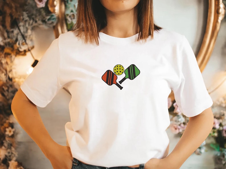 May include: White t-shirt featuring a colorful pickleball design. The graphic includes a yellow pickleball and two paddles, one red and one green. The paddles have black handles and vertical stripes.