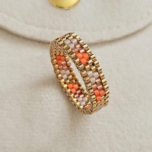 May include: A beaded ring with a gold-colored band and a pattern of orange, white, and gold beads. The ring is set against a white pouch with a gold snap closure.