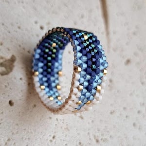 May include: A ring crafted from small, woven beads in a gradient pattern. The design features shades of dark navy, medium blue, and light blue, transitioning to white at the base. Small, metallic gold-colored beads are interspersed throughout the pattern.