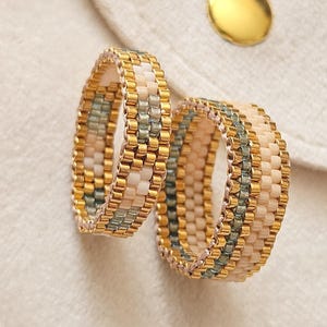 Puede incluir: Dos anillos elaborados con pequeñas cuentas cilíndricas en un patrón repetitivo de oro metálico, crema suave y verde salvia apagado. Las cuentas están dispuestas en un diseño tejido y estructurado, creando una banda de ancho constante.
